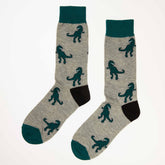 Et par lysegrå sokker med svarte hæler, mørkegrønne tupper og mansjetter, dekorert med gjentatte mønstre av en mørkegrønn T-Rex. Sokkene ligger flatt på en hvit bakgrunn.